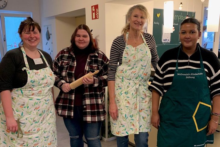 Backen für den guten Zweck Vier Frauen in Schürzen stehen in einer hellen Küche, bereit zum Backen, mit einem Nudelholz in der Hand.